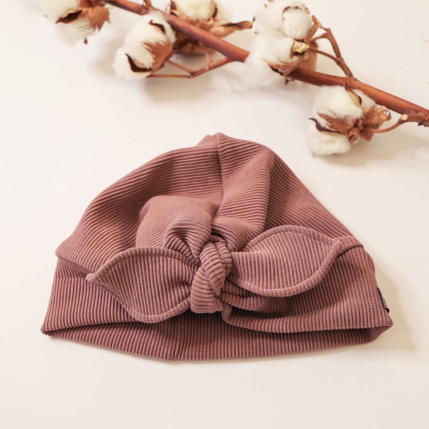 Altrosa turbanmütze mit Schleife aus geripptem Stoff, liegend vor Baumwollzweigen.