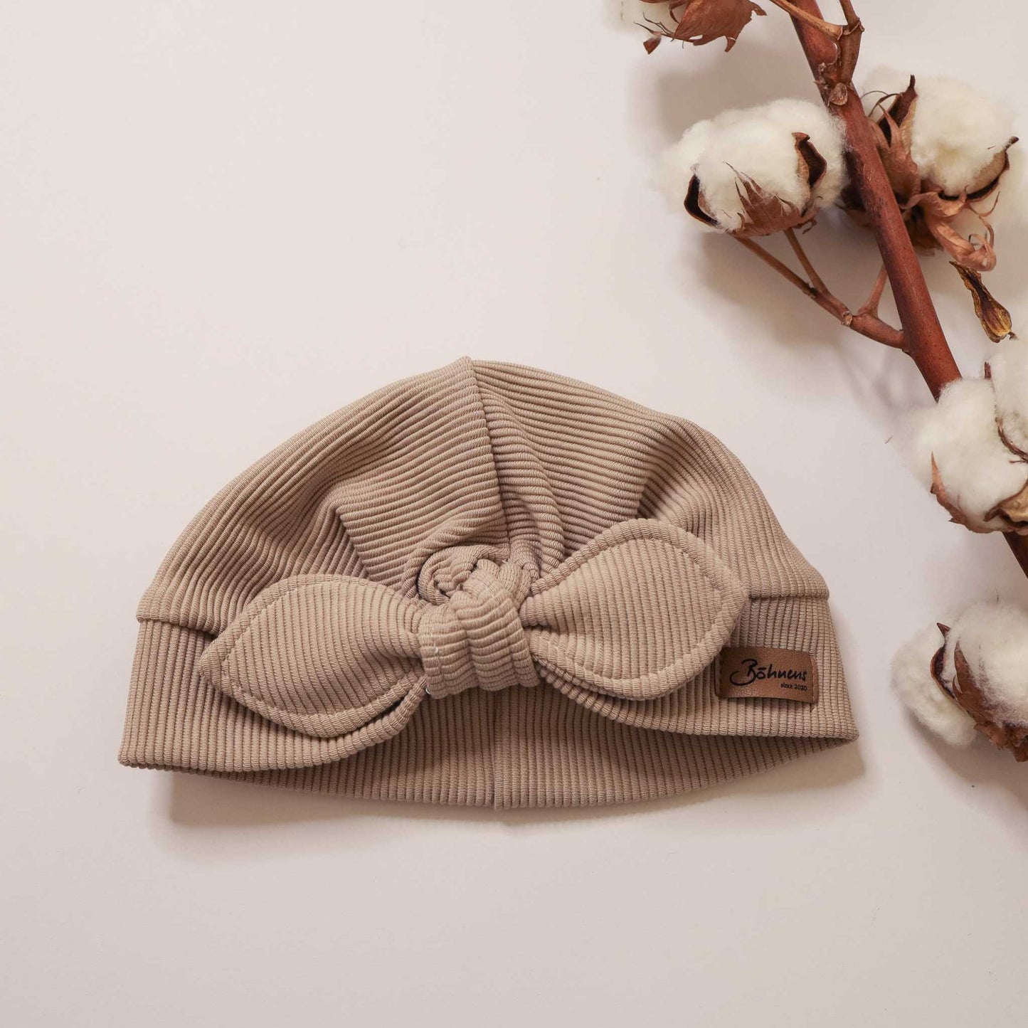 Dark beige turban hat with ribbed fabric and knot design, styled with cotton branches.