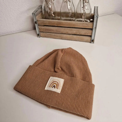 Regenbogen Beanie Größe 38 in braun mit Regenbogen-Aufnäher auf Tisch.