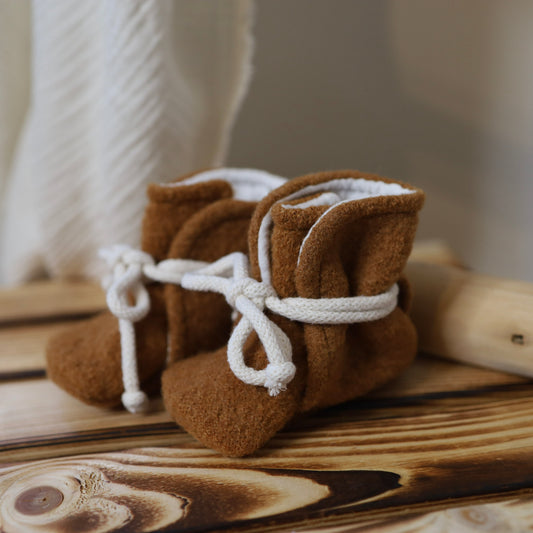 Handgenähte Babyschuhe aus braunem Wollwalk in Camel mit weissen Bindebändern auf einem Holzhintergrund